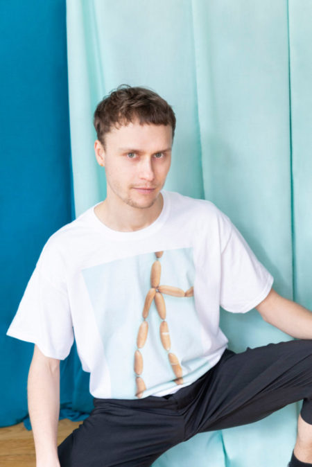 Ein Mann hockt vor blauem Hintergrund und trägt ein T-Shirt mit einer Würstchen-Kette drauf.