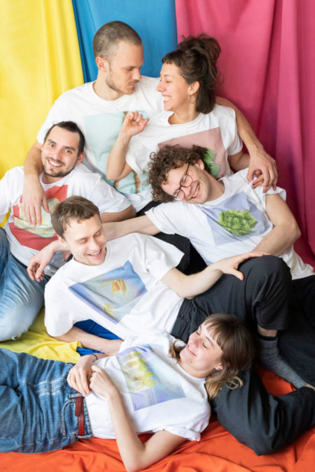 Sechs Personen sitzen vor einem bunten Hintergrund und tragen T-Shirts mit bunten Motiven.
